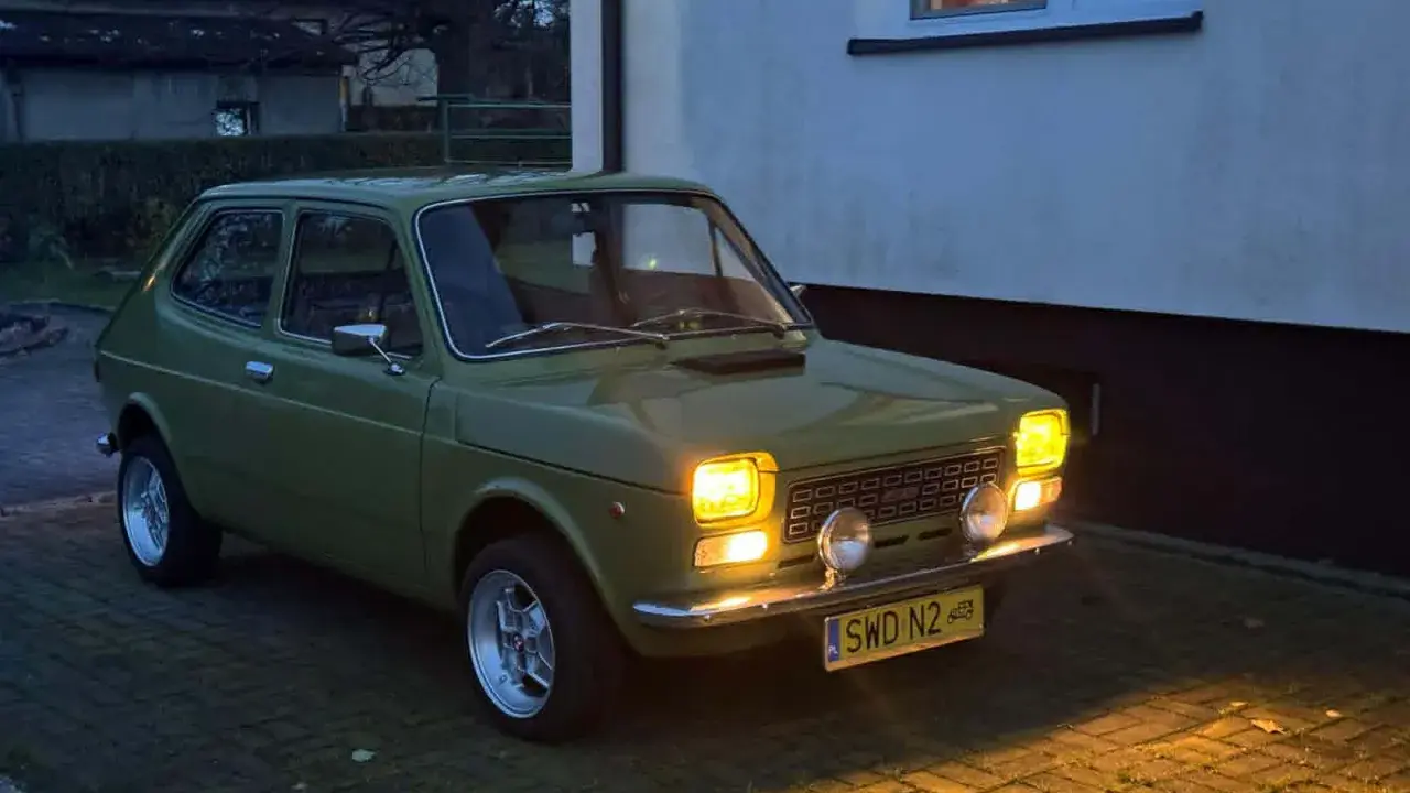 Zielony Fiat 127 z lat 70. stoi na podjeździe, jego światła rozświetlają zmierzch.