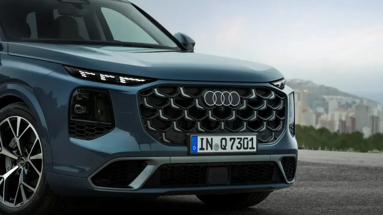 Przód eleganckiego Audi Q7 3.0 TDI w kolorze granatowym, z charakterystycznym grillem i światłami LED, na tle miejskiego krajobrazu.