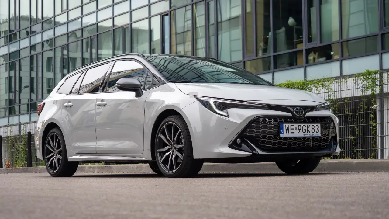 Biała Toyota Corolla 2.0 Hybrid kombi zaparkowana przed nowoczesnym budynkiem z lustrzaną fasadą.
