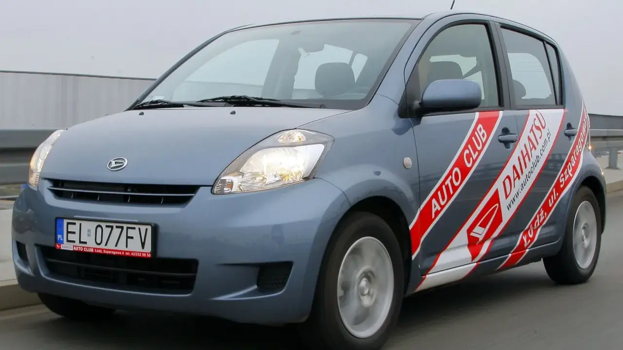 Niebieski Daihatsu Sirion z naklejkami "Auto Club Daihatsu" na drzwiach, poruszający się po drodze.