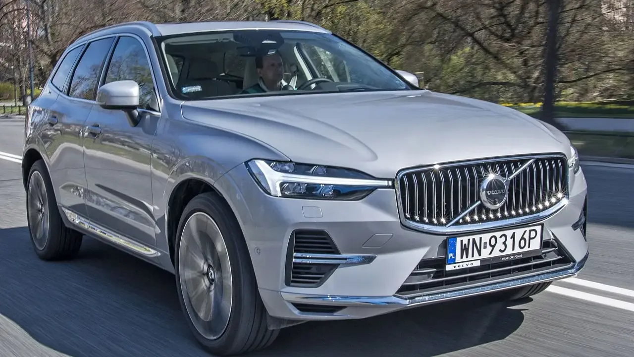 Srebrny Volvo XC60 2025 w ruchu, z kierowcą za kierownicą, na tle drzew.