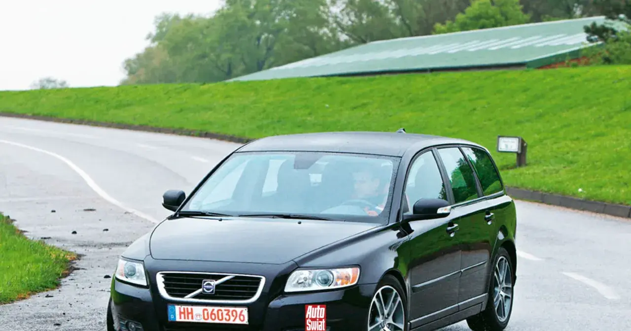 Czarne Volvo V50 2.0D na mokrej drodze, za kierownicą siedzi mężczyzna. W tle zielone wzgórza i budynek.