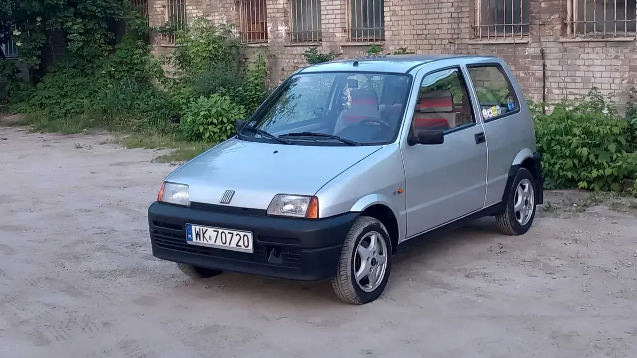 Srebrne cinquecento z czerwonymi akcentami we wnętrzu, zaparkowane na piaszczystym podwórku przed ceglanym budynkiem.