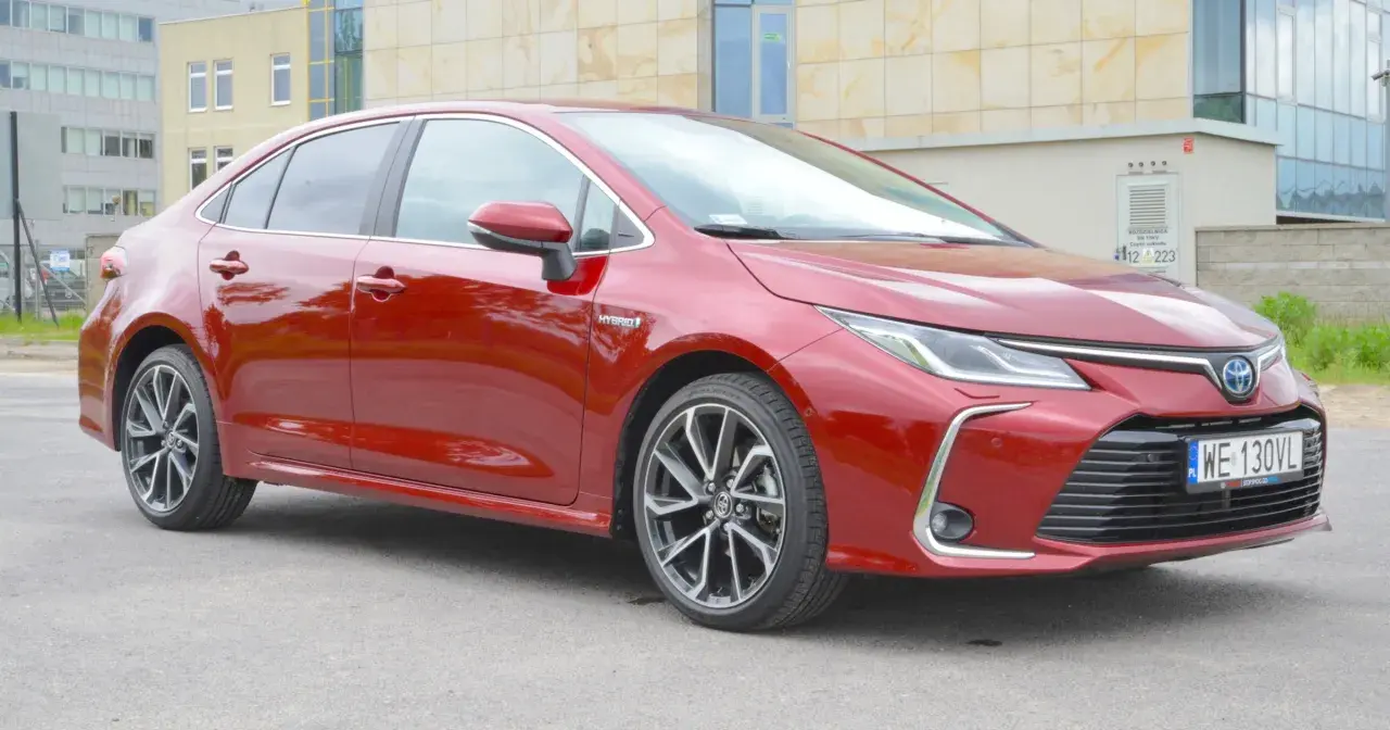 Błyszcząca, czerwona Toyota Corolla 1.8 Hybrid stoi na tle nowoczesnego budynku.