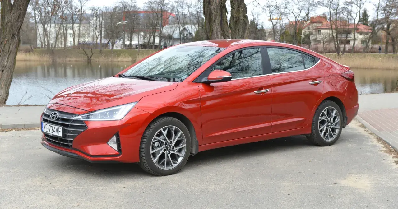 Czerwony Hyundai Elantra 2023 zaparkowany nad jeziorem, z drzewami i budynkami w tle.