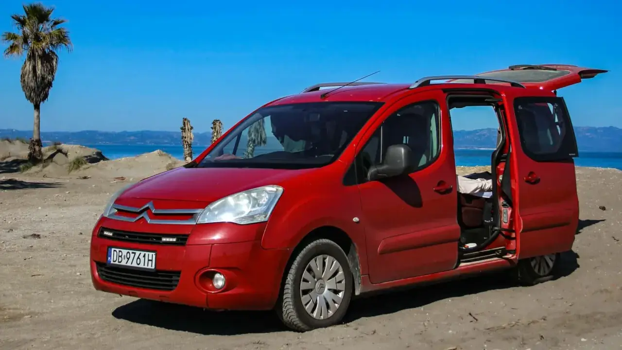 Czerwony Citroen Berlingo 1.6 HDI na plaży, z otwartymi drzwiami i bagażnikiem, na tle morza i palmy.