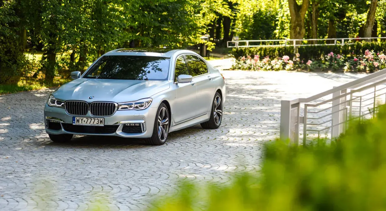 Srebrne BMW 750i lśni w słońcu na brukowanym podjeździe, otoczone zielenią i różami.