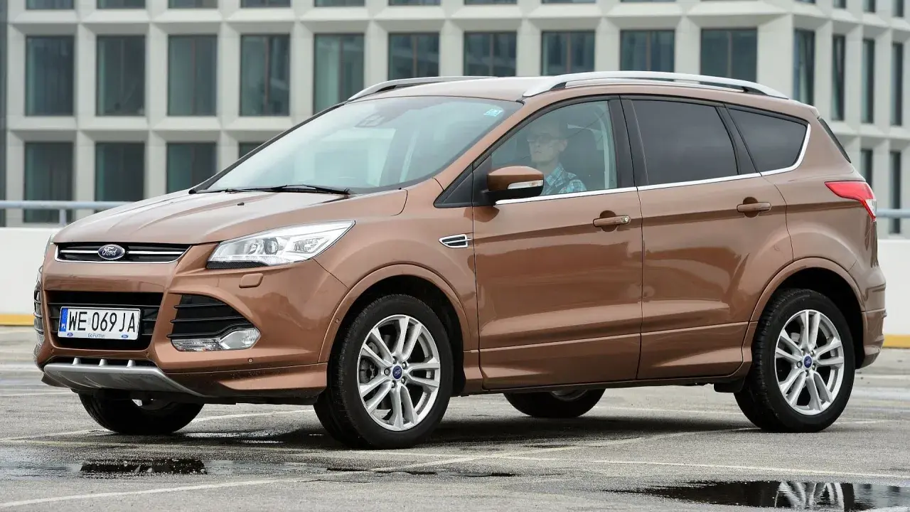 Brązowy Ford Kuga 2017 z kierowcą w okularach, zaparkowany na mokrym parkingu przed nowoczesnym budynkiem.