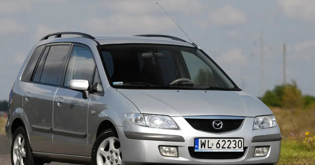 Srebrna Mazda Premacy z relingami dachowymi, zaparkowana na tle nieba i pól.