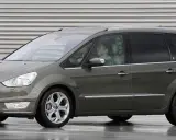 Szary Ford Galaxy MK2 z kierowcą za kierownicą, zaparkowany na mokrej nawierzchni przed budynkiem z poziomymi żaluzjami.