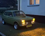 Zielony Fiat 127 z lat 70. stoi na podjeździe, jego światła rozświetlają zmierzch.