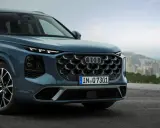 Przód eleganckiego Audi Q7 3.0 TDI w kolorze granatowym, z charakterystycznym grillem i światłami LED, na tle miejskiego krajobrazu.
