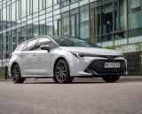 Biała Toyota Corolla 2.0 Hybrid kombi zaparkowana przed nowoczesnym budynkiem z lustrzaną fasadą.