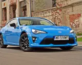 Błękitna Toyota GT86, znana też jako Toyota BRZ, zaparkowana przed zniszczonym budynkiem.