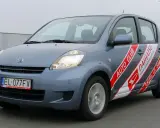 Niebieski Daihatsu Sirion z naklejkami "Auto Club Daihatsu" na drzwiach, poruszający się po drodze.