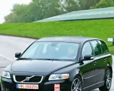 Czarne Volvo V50 2.0D na mokrej drodze, za kierownicą siedzi mężczyzna. W tle zielone wzgórza i budynek.