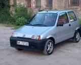 Srebrne cinquecento z czerwonymi akcentami we wnętrzu, zaparkowane na piaszczystym podwórku przed ceglanym budynkiem.