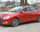 Czerwony Hyundai Elantra 2023 zaparkowany nad jeziorem, z drzewami i budynkami w tle.