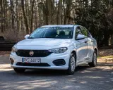 Biały Fiat Tipo 1.4 zaparkowany na leśnej drodze, otoczony drzewami i stertą drewna.