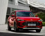 Czerwony MG ZS HEV, nowoczesny SUV hybrydowy, zaparkowany przed futurystycznym budynkiem.