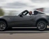 Mężczyzna w szarej kurtce prowadzi ciemnoszarą Mazdę Miata z opuszczonym dachem, jadąc po drodze.