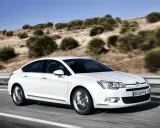 Biały Citroen C5 2.0 HDI pędzi po pustej drodze, otoczony suchymi, pagórkowatymi krajobrazami.