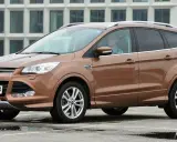 Brązowy Ford Kuga 2017 z kierowcą w okularach, zaparkowany na mokrym parkingu przed nowoczesnym budynkiem.