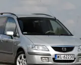 Srebrna Mazda Premacy z relingami dachowymi, zaparkowana na tle nieba i pól.