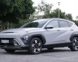 Nowy Hyundai Kona Hybrid w kolorze jasnoszarym, zaparkowany na brukowanej nawierzchni, otoczony zielenią.