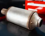 Metalowy tłumik, element układu wydechowego, leży na ciemnym tle. W tle widać fragment czerwonej skrzynki narzędziowej.