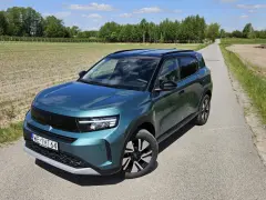 Nowy Opel Frontera 2025 w turkusowym kolorze na wiejskiej drodze, otoczony polami i drzewami.