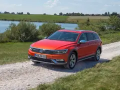 Pomarańczowy Volkswagen Passat Alltrack jedzie po żwirowej drodze, z jeziorem i zielonymi wzgórzami w tle.
