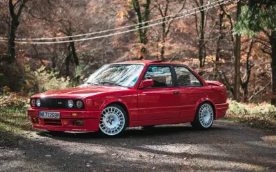 Czerwone bmw e30 coupe z białymi felgami na tle jesiennego lasu.