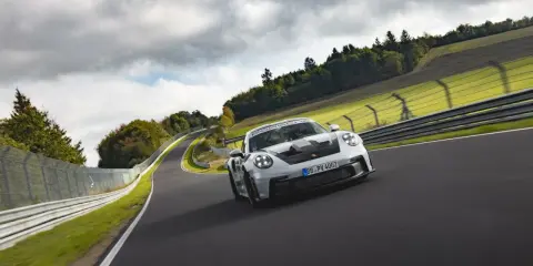 Biały Porsche 911 GT3 RS z czarnymi akcentami pędzi po torze wyścigowym wśród zielonych wzgórz i chmur.