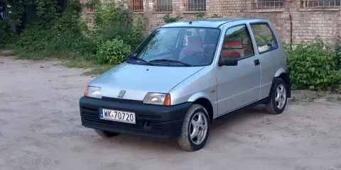Srebrne cinquecento z czerwonymi akcentami we wnętrzu, zaparkowane na piaszczystym podwórku przed ceglanym budynkiem.