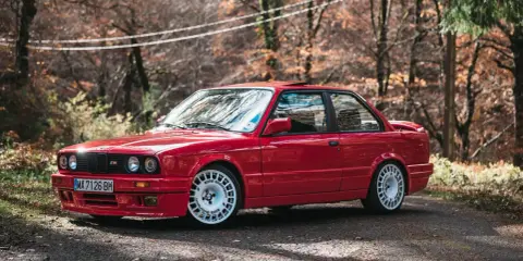 Czerwone bmw e30 coupe z białymi felgami na tle jesiennego lasu.