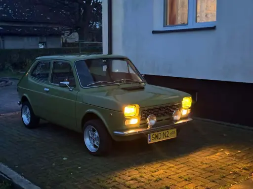 Zielony Fiat 127 z lat 70. stoi na podjeździe, jego światła rozświetlają zmierzch.