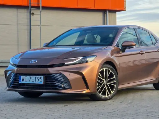 Brązowa Toyota Camry 2.5 Hybrid z eleganckimi felgami stoi na tle nowoczesnego budynku.