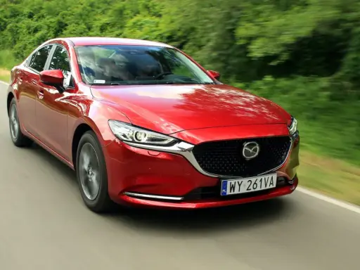 Czerwona Mazda 6 2.0 benzyna pędzi po wiejskiej drodze, otoczona zielenią drzew.