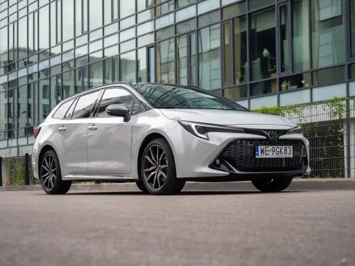 Biała Toyota Corolla 2.0 Hybrid kombi zaparkowana przed nowoczesnym budynkiem z lustrzaną fasadą.