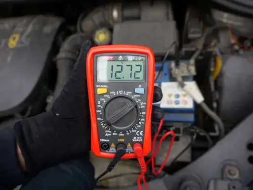 Ręka w czarnej rękawiczce trzyma multimetr pokazujący 12.72V, co pomaga sprawdzić akumulator.
