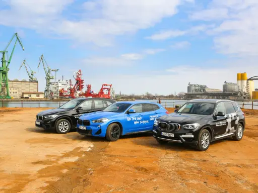 Trzy SUV-y BMW stoją na placu budowy, w tle portowe dźwigi i budynki.