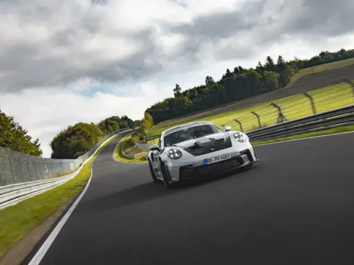 Biały Porsche 911 GT3 RS z czarnymi akcentami pędzi po torze wyścigowym wśród zielonych wzgórz i chmur.