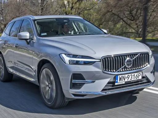Srebrny Volvo XC60 2025 w ruchu, z kierowcą za kierownicą, na tle drzew.