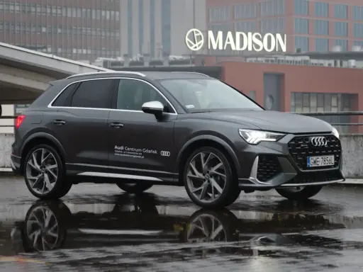 Szary Audi RS Q3 na mokrej nawierzchni, z odbiciem w kałuży. W tle budynek z napisem MADISON.