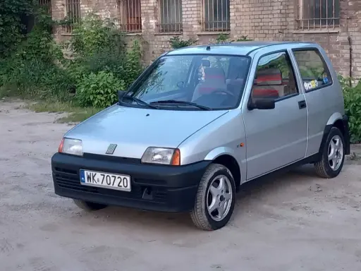 Srebrne cinquecento z czerwonymi akcentami we wnętrzu, zaparkowane na piaszczystym podwórku przed ceglanym budynkiem.