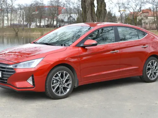 Czerwony Hyundai Elantra 2023 zaparkowany nad jeziorem, z drzewami i budynkami w tle.