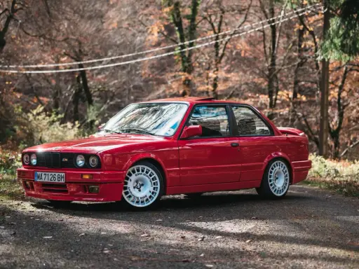 Czerwone bmw e30 coupe z białymi felgami na tle jesiennego lasu.