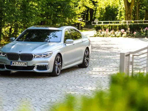 Srebrne BMW 750i lśni w słońcu na brukowanym podjeździe, otoczone zielenią i różami.