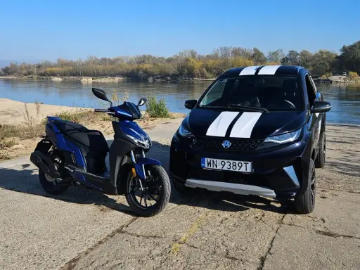 Niebieski skuter i czarne aixam auto z białymi pasami stoją nad rzeką.